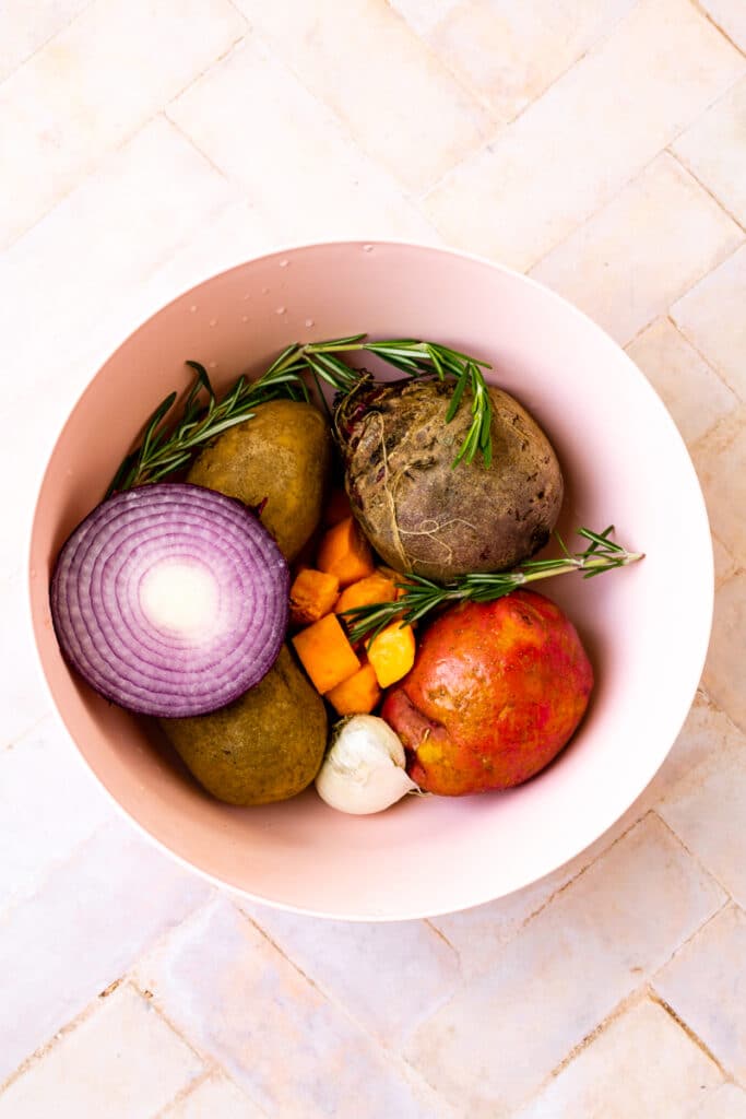 pink bowl with red onion, potatoes, sweet potatoes, garlic, and rosemary